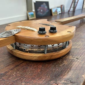 May include: A unique, handmade wooden banjo-guitar hybrid instrument. The body features a light brown wood construction with a circular metal resonator. It has a wooden neck with strings, tuning knobs, and a decorative metal tailpiece. The instrument rests on a wooden table.