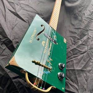 May include: A handmade green cigar box guitar with the text "H. UPMANN" and "THE BANKER BAIT TRADER". It features a wooden neck, brass hardware, and black control knobs. The strings have colourful accents.
