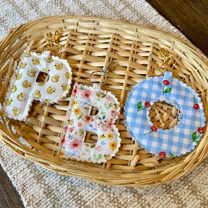 May include: Three fabric letter keychains with gold-tone metal clasps. The letter P features a yellow duck pattern, the letter B has a colorful floral print, and the letter O displays a blue and white gingham pattern with red cherries.