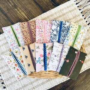 May include: A collection of ten rectangular fabric tissue holders featuring various patterns including floral prints, bees, strawberries, mushrooms, and blue toile, each with a contrasting fabric center strip, arranged in a woven wicker basket.