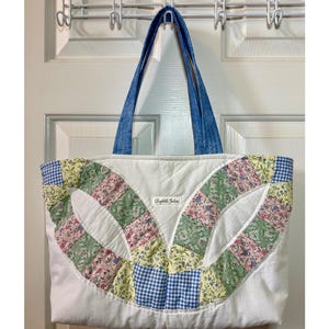 May include: A quilted tote bag featuring a white base with a patchwork design of floral and blue gingham fabric patterns. The bag has two blue denim-style handles and a small white rectangular label with the text Elizabeth Julia centered.