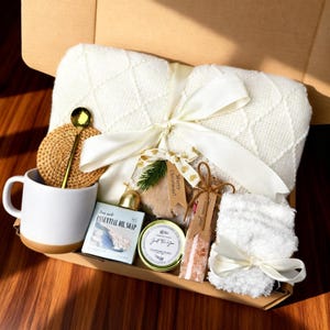 May include: A gift box filled with a white knitted blanket tied with a cream ribbon, a white mug with a brown base, a gold spoon, a woven coaster, essential oil soap, a candle, bath salts, and a fluffy white washcloth. The box includes a tag that says "Merry Christmas!"