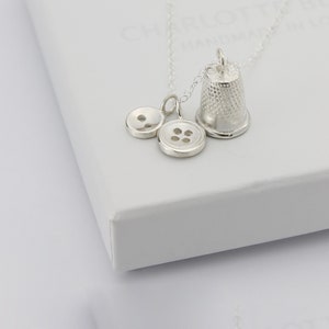 May include: Silver necklace with a thimble charm and two button charms. The necklace is displayed on a white box.