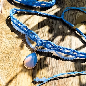 Può includere: Una collezione di gioielli in macramè blu, tra cui una collana con un pendente a goccia, un braccialetto e un filo di perline. Il pendente ha una montatura color oro e una pietra azzurra e rosa. I gioielli sono esposti su una superficie di legno.