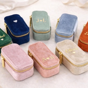 May include: A selection of velvet ring boxes in shades like green, blue, and pink. Each box features a gold zip and personalised text or initials. The rectangular boxes are designed for storing jewellery.