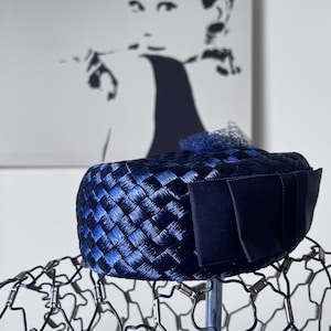 Puede incluir: Un sombrero tejido azul marino con un diseño estructurado y un lazo azul oscuro. El sombrero tiene una apariencia texturizada y un pequeño acento decorativo en la parte superior. El fondo presenta una imagen en blanco y negro de una mujer.