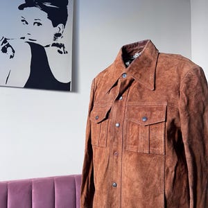 May include: A brown suede jacket with a collared neckline and two chest pockets. The jacket has button closures and a classic design. The image also shows a black and white portrait of a woman on the wall.