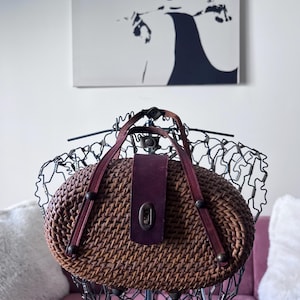 以下が含まれることがあります： 楕円形の茶色の織りハンドバッグ。バーガンディ色の革のフラップとハンドルが付いています。バッグには金属製の留め金があり、ワイヤーマネキンに展示されています。背景には白黒の肖像画があります。