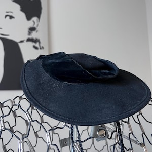 May include: A dark navy blue hat with a rounded crown and a wide brim. The hat is made of a soft, velvety material and is displayed on a wire mannequin head. A black and white portrait is visible in the background.