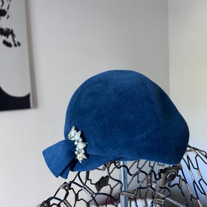 Puede incluir: Un sombrero cloche azul marino con un ala pequeña y un adorno de joyas decorativo. El sombrero está hecho de un material suave y aterciopelado y se exhibe en una cabeza de maniquí de alambre.