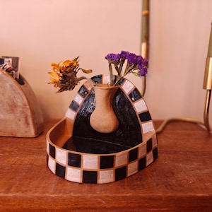 Pode incluir: Um vaso decorativo de cerâmica com um design xadrez em preto e branco, contendo flores amarelas e roxas secas. O vaso tem uma forma única e está sobre uma superfície de madeira.