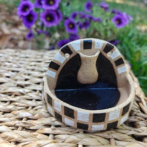 May include: A small, handmade ceramic fountain with a black interior and a beige vase-shaped element. The rim is decorated with a checkerboard pattern of black and white squares. The fountain sits on a woven surface, with purple flowers in the background.