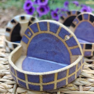 Könnte beinhalten: Handgemachte Keramik-Seifenschale mit einzigartigem Design. Die Schale hat einen tiefvioletten Innenraum und einen Rand mit einem Gittermuster in Lila und Gold. Die Schale steht vor einem natürlichen, gewebten Hintergrund mit lila Blumen im Hintergrund.