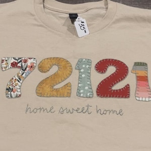 Op de afbeelding: Beige t-shirt met de cijfers 72121 op de voorkant. De 7 is bloemen, de 2 is geel, de 1 is lichtblauw met witte stippen, de andere 2 is rood en de laatste 1 is gestreept. De woorden "home sweet home" zijn eronder geborduurd.