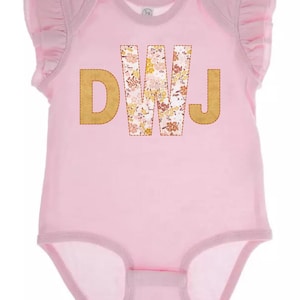 May include: A light pink baby onesie with ruffled sleeves. The onesie features the letters "DWJ" in a gold color, with the "W" filled with a floral pattern. The onesie has a snap closure at the bottom.