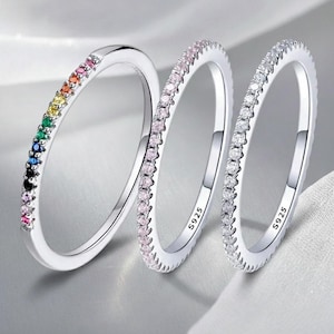 Op de afbeelding: Drie zilveren ringen met verschillende edelsteenarrangementen. Eén ring heeft een regenboog van gekleurde edelstenen, de andere roze en heldere edelstenen. Elke ring is gemarkeerd met "S925".