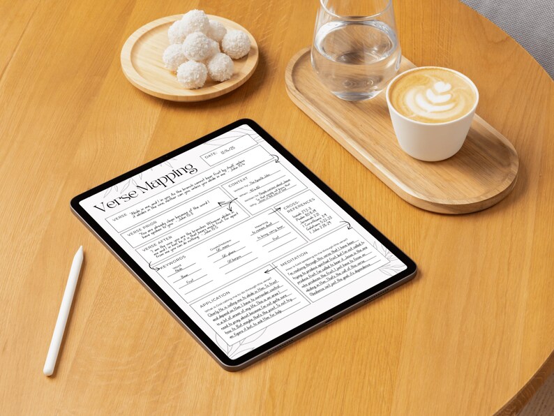 May include: A digital tablet displaying a "Verse Mapping" worksheet, alongside a white stylus. A wooden tray holds a glass of water and a cup of coffee with latte art. A small wooden plate with white, round treats is also present.
