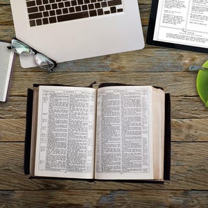 May include: An open book with text, a laptop, a tablet, a cup of coffee, a notebook, and glasses on a wooden surface. The book is open to a page of text. The laptop is partially visible. The tablet displays text. The coffee is in a green mug.