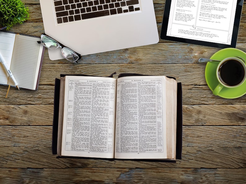 May include: An open book with text, a laptop, a tablet, a cup of coffee, a notebook, and glasses on a wooden surface. The book is open to a page of text. The laptop is partially visible. The tablet displays text. The coffee is in a green mug.