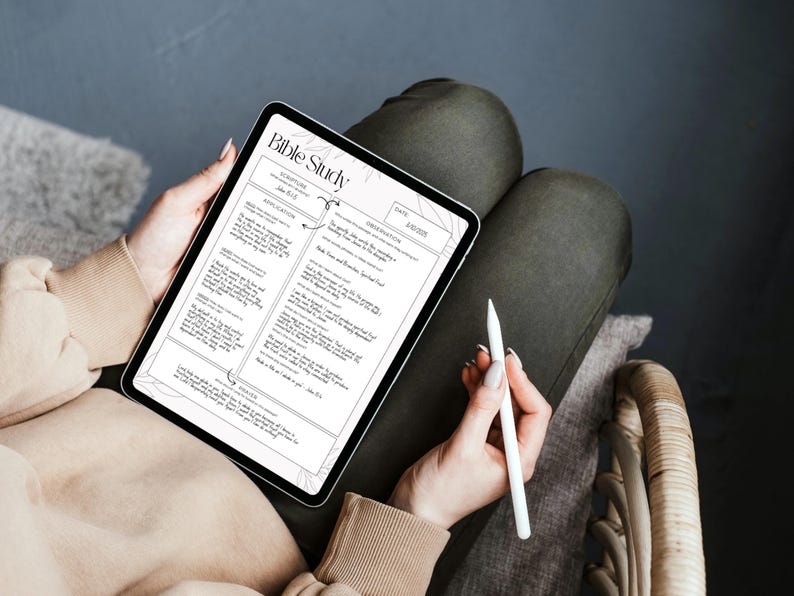May include: A digital tablet displaying a "Bible Study" worksheet with text and diagrams. A white stylus rests in the hand of a person holding the tablet. The person is wearing a beige sweater and dark green pants.