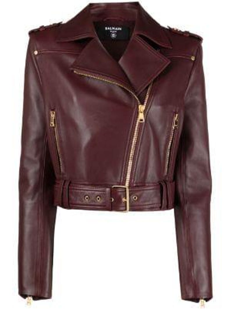 May include: A burgundy leather motorcycle jacket with a notched collar, asymmetrical zip closure, and a belted waist. The jacket features gold-tone hardware, including zippers and belt buckle. The sleeves have zip cuffs and shoulder epaulettes.