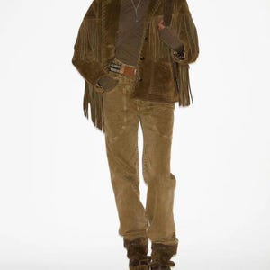 May include: A full-length shot of a person wearing a brown suede jacket with fringe, a brown turtleneck, corduroy pants, and brown suede boots. The jacket has a button closure and a belt. The outfit is a monochromatic ensemble.