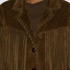 May include: A brown suede jacket with fringe detailing along the chest and sleeves. The jacket features decorative stitching and two dark brown buttons. The collar is notched, and the jacket appears to be a medium weight.