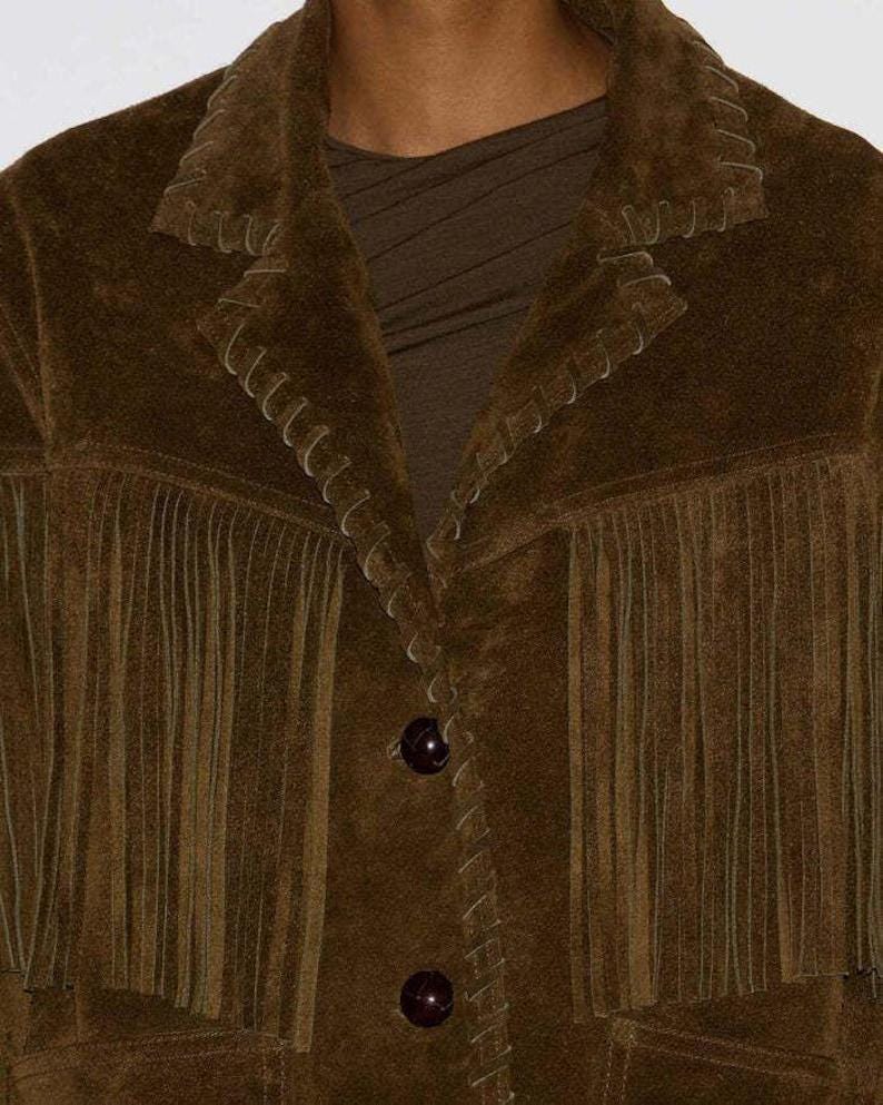 May include: A brown suede jacket with fringe detailing along the chest and sleeves. The jacket features decorative stitching and two dark brown buttons. The collar is notched, and the jacket appears to be a medium weight.