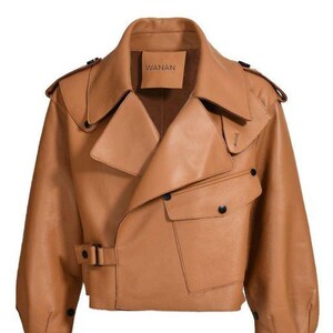May include: A tan leather jacket with a wide collar, asymmetrical front, and two front pockets. The jacket has a belt at the waist and the brand name "WANAN" is visible on a tag. The sleeves are long and the jacket has black buttons.
