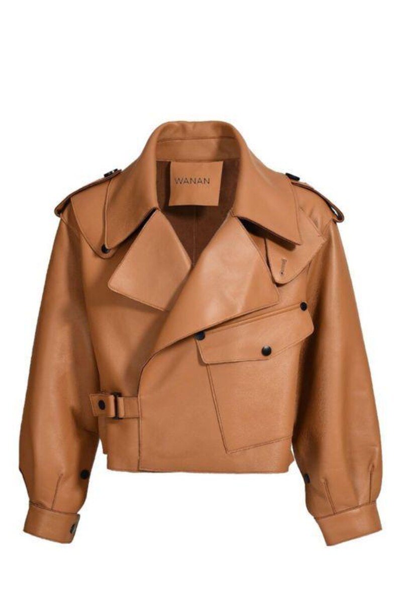 May include: A tan leather jacket with a wide collar, asymmetrical front, and two front pockets. The jacket has a belt at the waist and the brand name "WANAN" is visible on a tag. The sleeves are long and the jacket has black buttons.