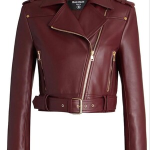 May include: A burgundy leather motorcycle jacket with a notched collar, asymmetrical zipper, and gold-tone hardware. The jacket features a belted waist, shoulder epaulets, and multiple zippered pockets. The brand name "BALMAIN PARIS" is visible on a tag.