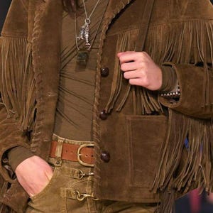 May include: A brown suede jacket with fringe detailing, paired with matching pants and a belt. The jacket features button closures and pockets. A patterned scarf and necklace complete the look. The outfit is a stylish take on Western wear.