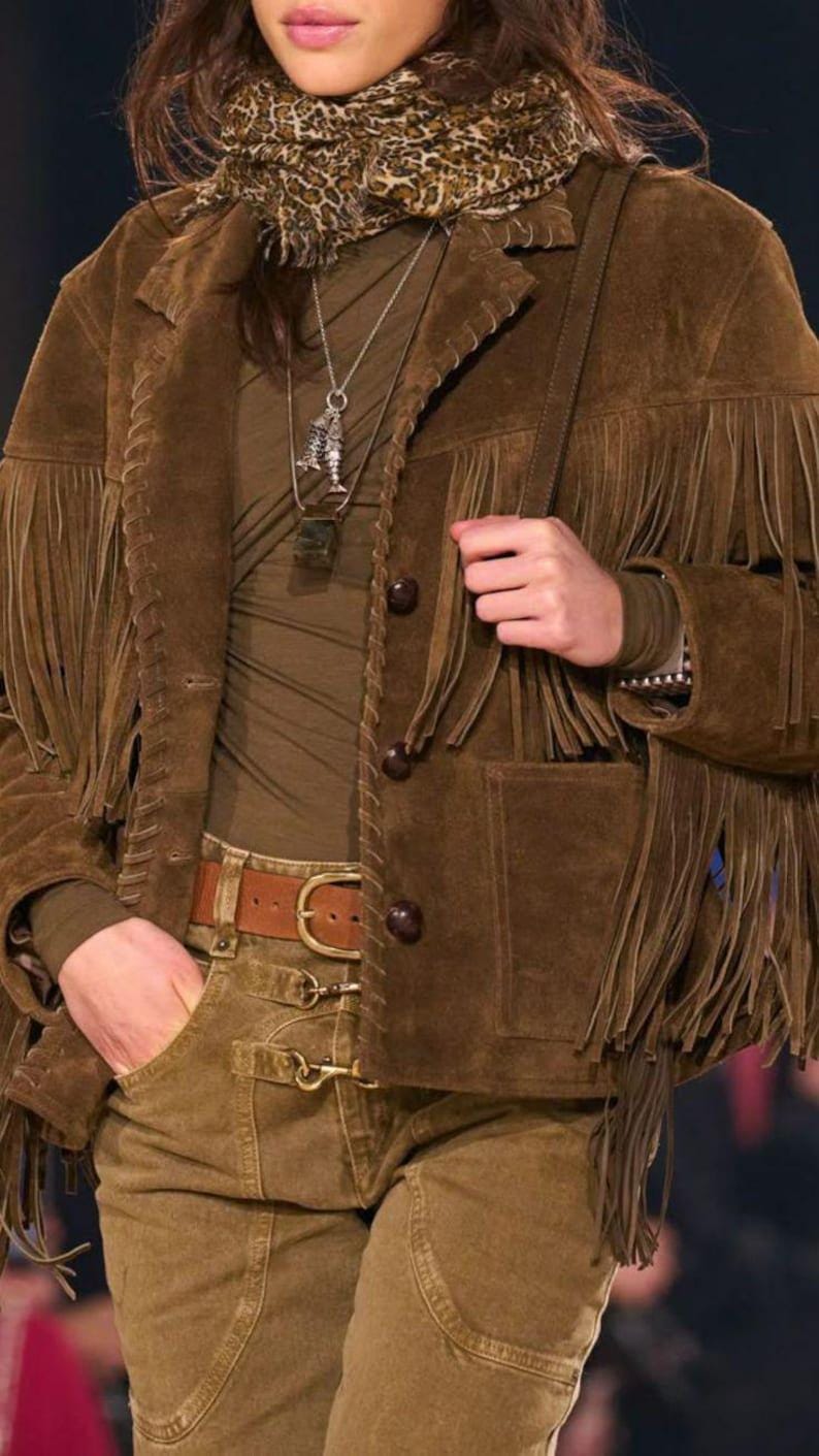 May include: A brown suede jacket with fringe detailing, paired with matching pants and a belt. The jacket features button closures and pockets. A patterned scarf and necklace complete the look. The outfit is a stylish take on Western wear.