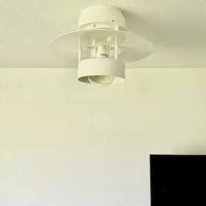 Może przedstawiać: Biała, nowoczesna lampa sufitowa z okrągłym kloszem i cylindryczną podstawą. Lampa ma duży, płaski, okrągły klosz i mniejszą, cylindryczną podstawę. Żarówka jest widoczna. Oprawa jest zamontowana na białym suficie.