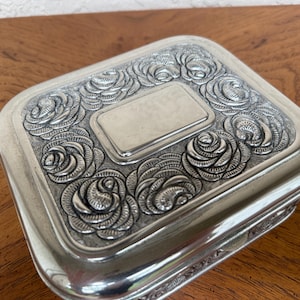 May include: A rectangular silver-toned metal box featuring a raised relief pattern of roses covering the lid. The center of the lid contains a smooth, blank rectangular plaque. The box has rounded corners and a polished, reflective metallic finish.