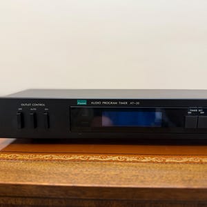 May include: Black Sansui Audio Program Timer AT-20. The device has a digital display and several control buttons. The front panel includes the text "OUTLET CONTROL" and "AUDIO PROGRAM TIMER AT-20". The timer is set on a wooden surface.