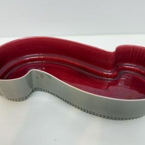 May include: A ceramic serving dish with a unique, elongated shape. The interior is a glossy, deep red, contrasting with the gray exterior. The rim has a textured, ridged design. This decorative dish is ideal for serving snacks or as a decorative piece.
