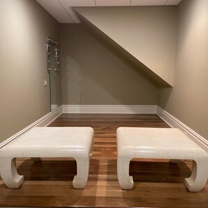 May include: Two square, off-white coffee tables with curved legs are positioned on a hardwood floor. The tables have a textured surface and are in a room with neutral-colored walls and a slanted ceiling.