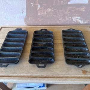 Può includere: Tre stampi in ghisa nera per pane a forma di pannocchia, ognuno con sette cavità individuali a forma di mais con texture a chicchi. Queste teglie rettangolari presentano manici integrati su entrambe le estremità e una finitura metallica scura.