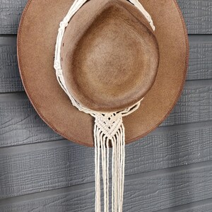 May include: A brown hat with a wide brim is suspended by a cream-coloured macrame hat hanger. The hanger features a wooden ring at the top and a decorative knot design.