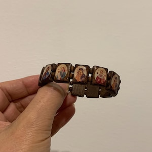 May include: A wooden bracelet featuring rectangular links with religious icons. Each link displays a different image, with a dark brown wood finish. The bracelet is held by a hand against a plain background.