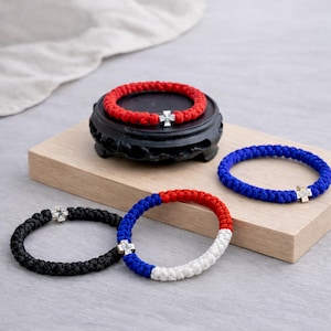 Könnte beinhalten: Eine Sammlung geflochtener Armbänder in verschiedenen Farben, darunter Rot, Blau, Schwarz und ein dreifarbiges Design. Jedes Armband hat einen silbernen Kreuzanhänger. Ein rotes Armband liegt auf einem schwarzen Dekorationsständer, andere auf einer Holzoberfläche.