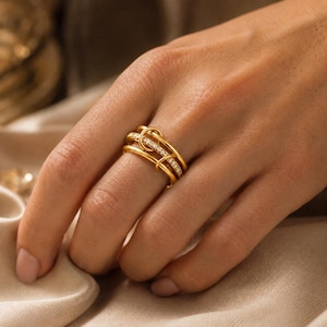 Peut inclure: Une bague dorée au design unique, composée de plusieurs anneaux. L'un des anneaux est orné de petites pierres scintillantes. La bague est présentée sur une main, mettant en valeur ses détails complexes et son style élégant.