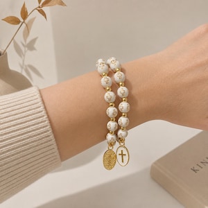 May include: White beaded bracelet with gold accents and cross charms. The bracelet features white beads with gold crosses and gold-colored spacers. Two charms hang from the bracelet, one with a cross and the other with a design.