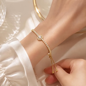 Könnte beinhalten: Ein verstellbares goldfarbenes Armband mit einem zentralen runden Anhänger, der mit einem klaren Stein besetzt und von kleineren Steinen umgeben ist, verbunden mit einer zarten Kette aus klaren Steinen und zwei goldenen Perlen am Verschluss.