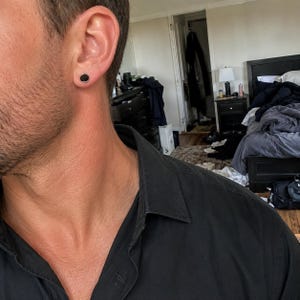 May include: A circular stud earring featuring a smooth, matte black center inlay surrounded by a thin, polished silver-tone metal rim. The earring is worn in the earlobe, presenting a simple, flat, and geometric design that sits flush against the skin.