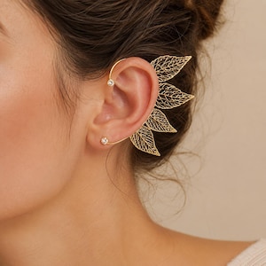 May include: A gold-toned ear cuff featuring a delicate filigree leaf design that extends along the outer edge of the ear. The piece includes two small, clear, round stones set at the top and bottom ends of the curved metal wire frame.