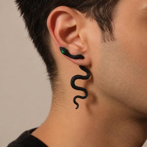 May include: A black snake-shaped earring featuring a textured, winding body that extends downward from the earlobe. The snake has a distinct head positioned at the top of the ear with a small, bright green eye detail visible on the side of the head.
