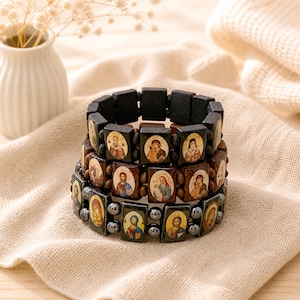 Puede incluir: Tres pulseras apiladas con iconos religiosos. La pulsera superior tiene bloques de madera negros. La pulsera del medio tiene bloques de madera marrones. La pulsera inferior tiene cuentas de color gris oscuro. Cada bloque y cuenta presenta un icono diferente con detalles dorados y coloridos.