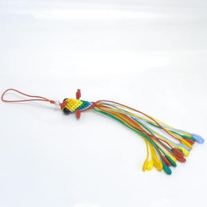 May include: A colorful, handmade fish-shaped charm. The charm is crocheted with yellow, green, blue, and red threads. It has black bead eyes and a long tail of multicolored strands. A small loop is attached for hanging.